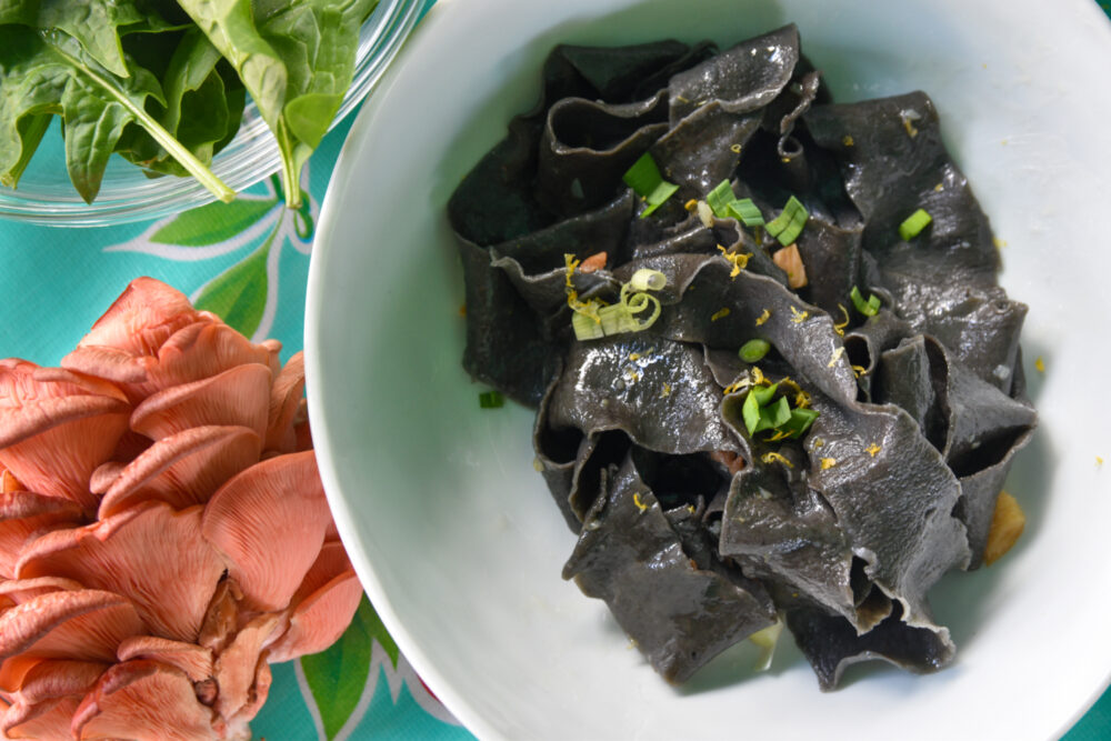 A bowl of black squid ink pasta, and spinach and pink oyster mushrooms on the side